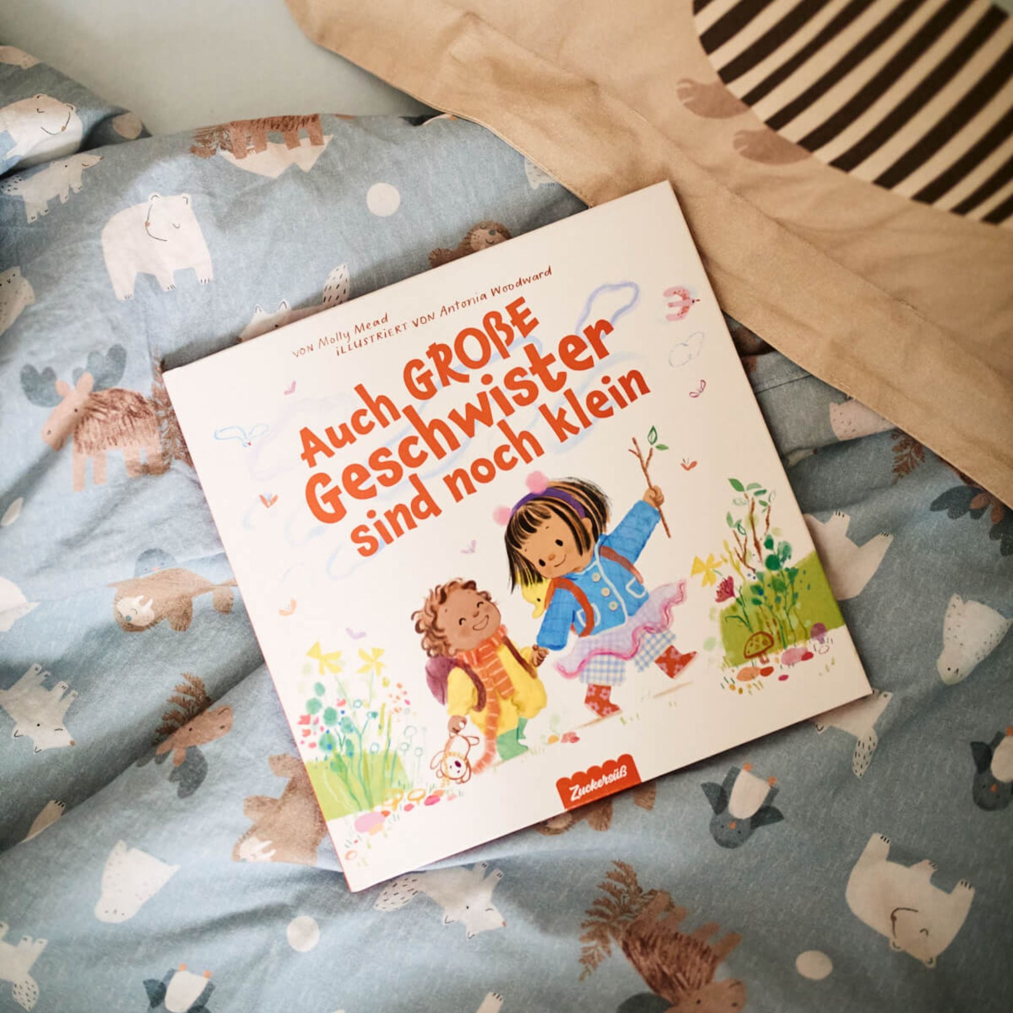 Das deutsche Bilderbuch "Auch große Geschwister sind noch klein" aus dem Zuckersüß Verlag liegt auf einer blauen Tagesdecke mit Tiermotiven; auf dem Cover sind ein Mädchen und ein Junge zu sehen, die draußen spielen und ihre Gefühle erkunden.