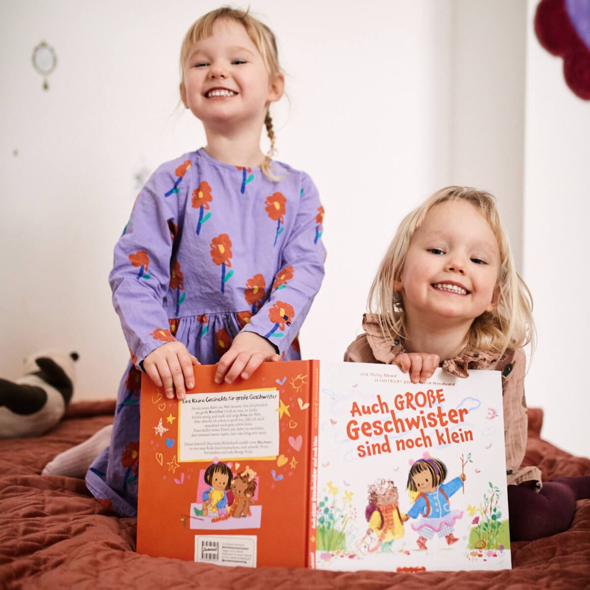 Zwei Kinder lächeln und halten das Buch "Auch große Geschwister sind noch klein" aus dem Zuckersüß Verlag in der Hand. Sie sitzen auf einem Bett inmitten von Stofftieren und erzählen sich Geschichten und Gefühle über das große Geschwisterdasein.