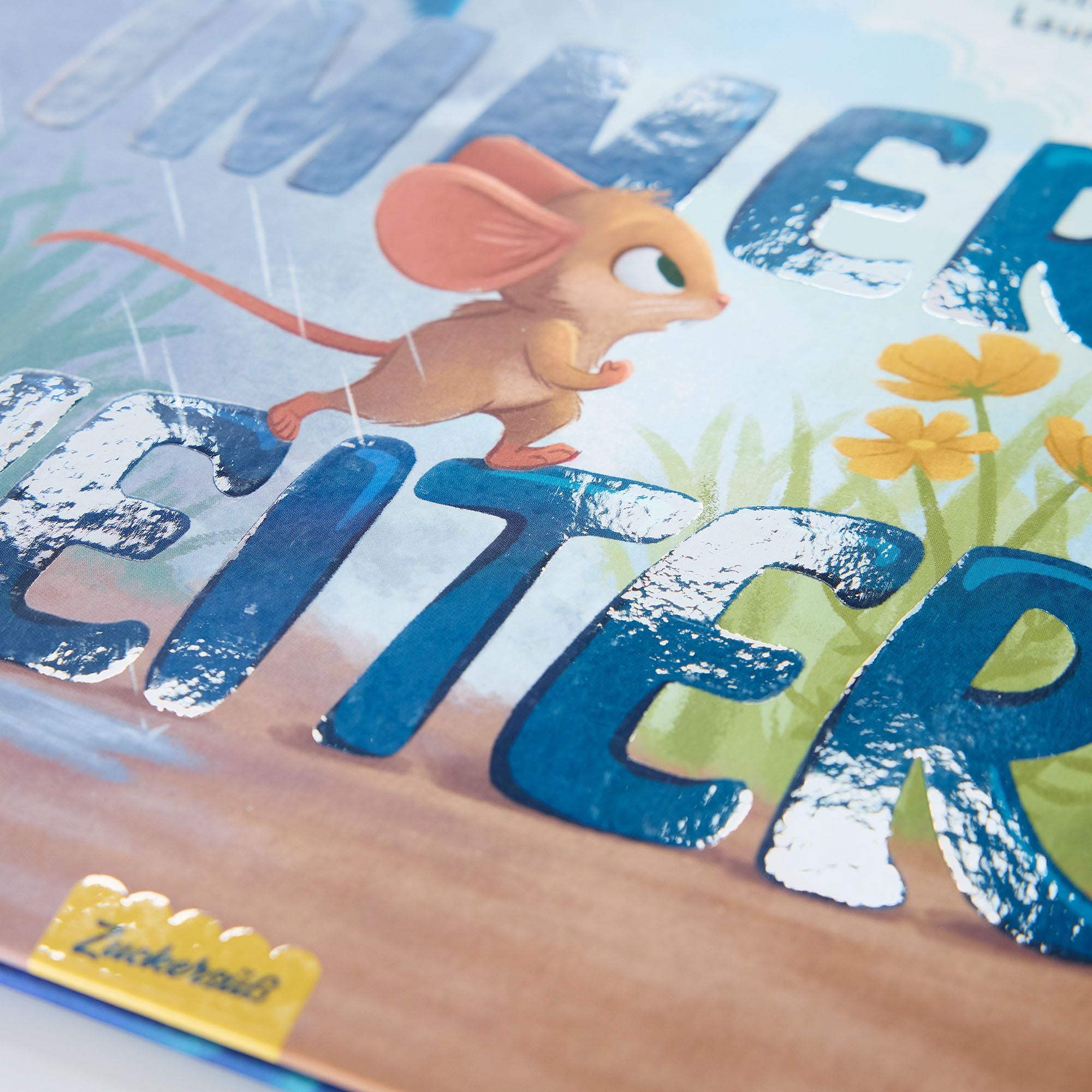 Eine Nahaufnahme des Umschlags des Mutmachbuchs "Immer weiter" des Zuckersüß Verlags zeigt eine illustrierte Maus, die an gelben Blumen, leuchtend blauen Buchstaben und einem gelben Etikett für Kinder mit Emotionen und Gefühlen vorbeiläuft.