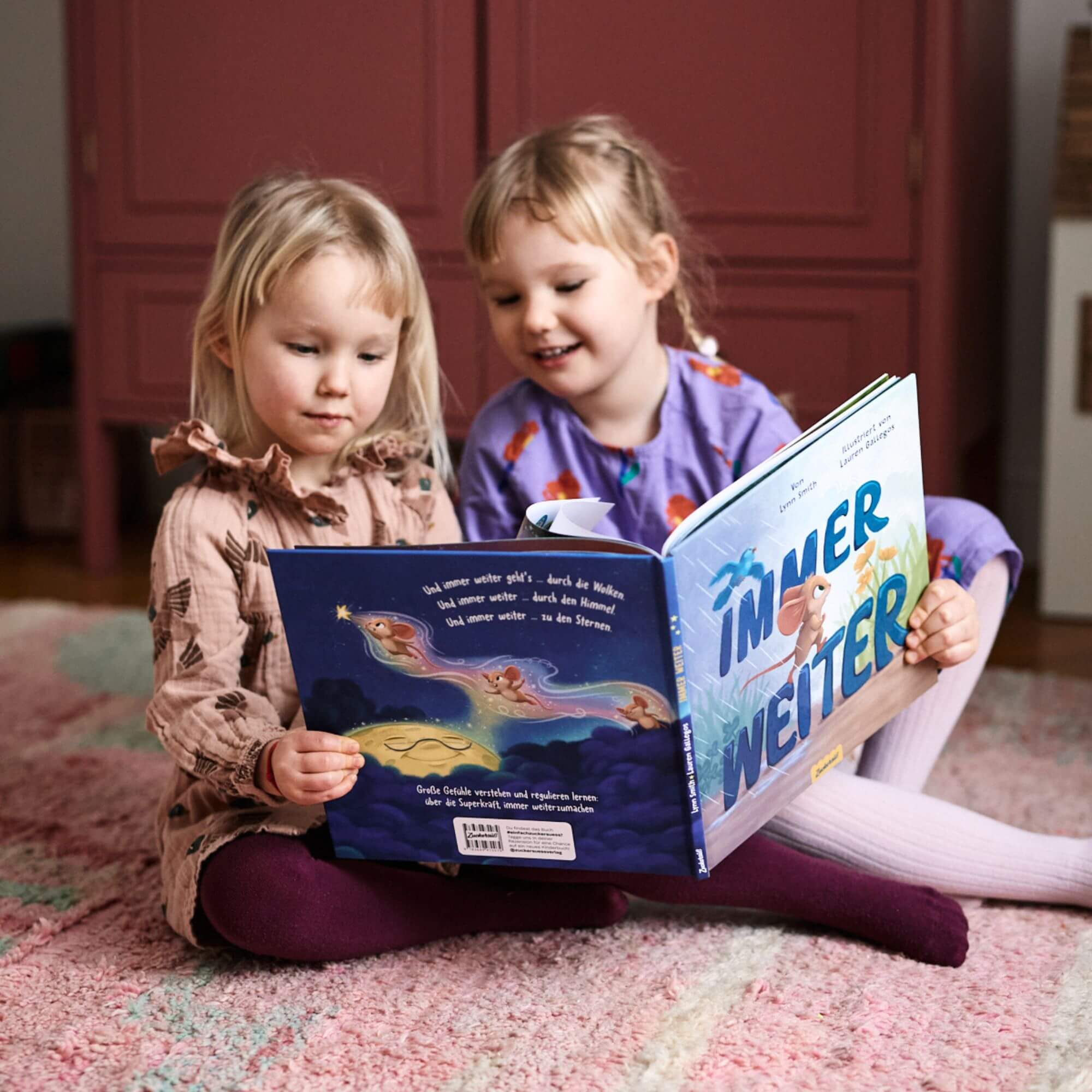 Zwei junge Mädchen sitzen auf dem Boden und lesen "Immer weiter" aus dem Zuckersüß Verlag. Die eine trägt ein hellbraunes Kleid, die andere ein lavendelfarbenes; sie lächeln herzlich und erzählen in ihrem gemütlichen Zimmer Geschichten über die Emotionen und Gefühle von Kindern.