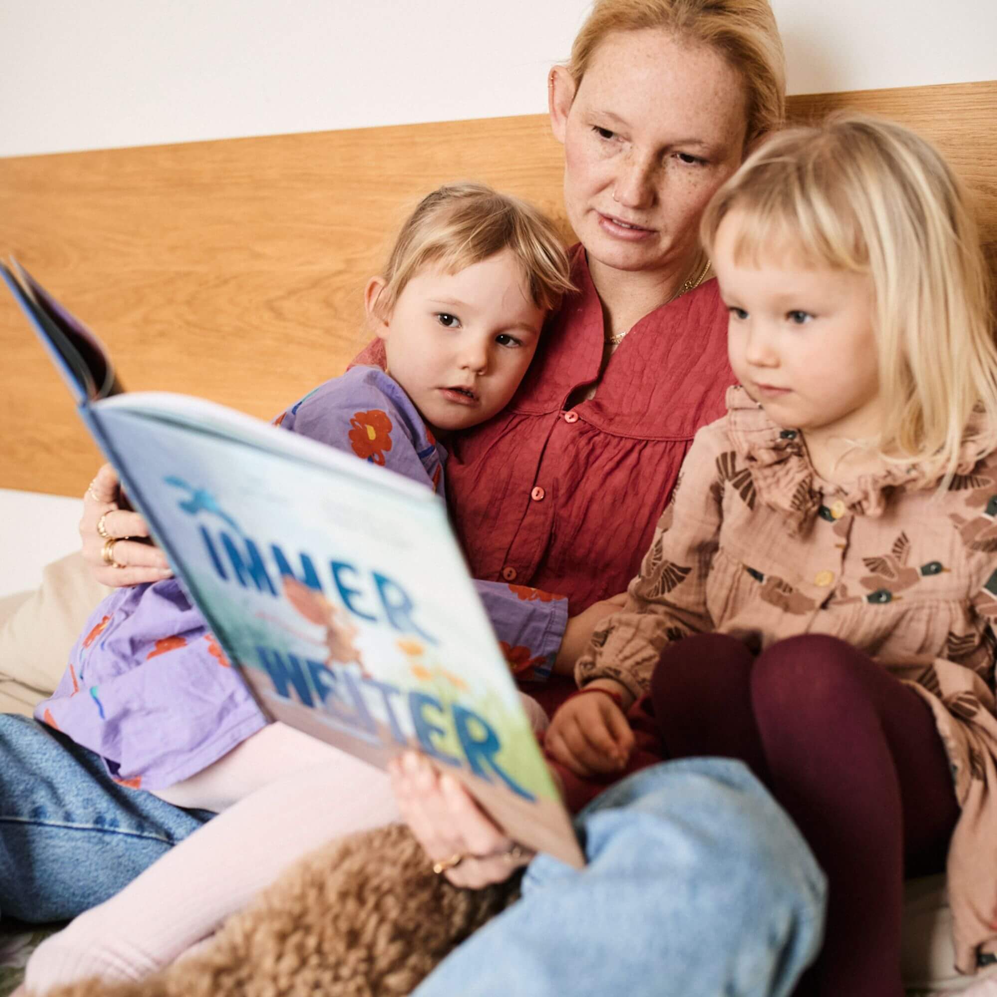 Eine Frau sitzt auf einem Bett und liest "Immer weiter" aus dem Zuckersüß Verlag zwei kleinen Kindern vor, die sich aneinander kuscheln und aufmerksam zuhören, während sie lernen, gemeinsam Emotionen bei Kindern zu verstehen.