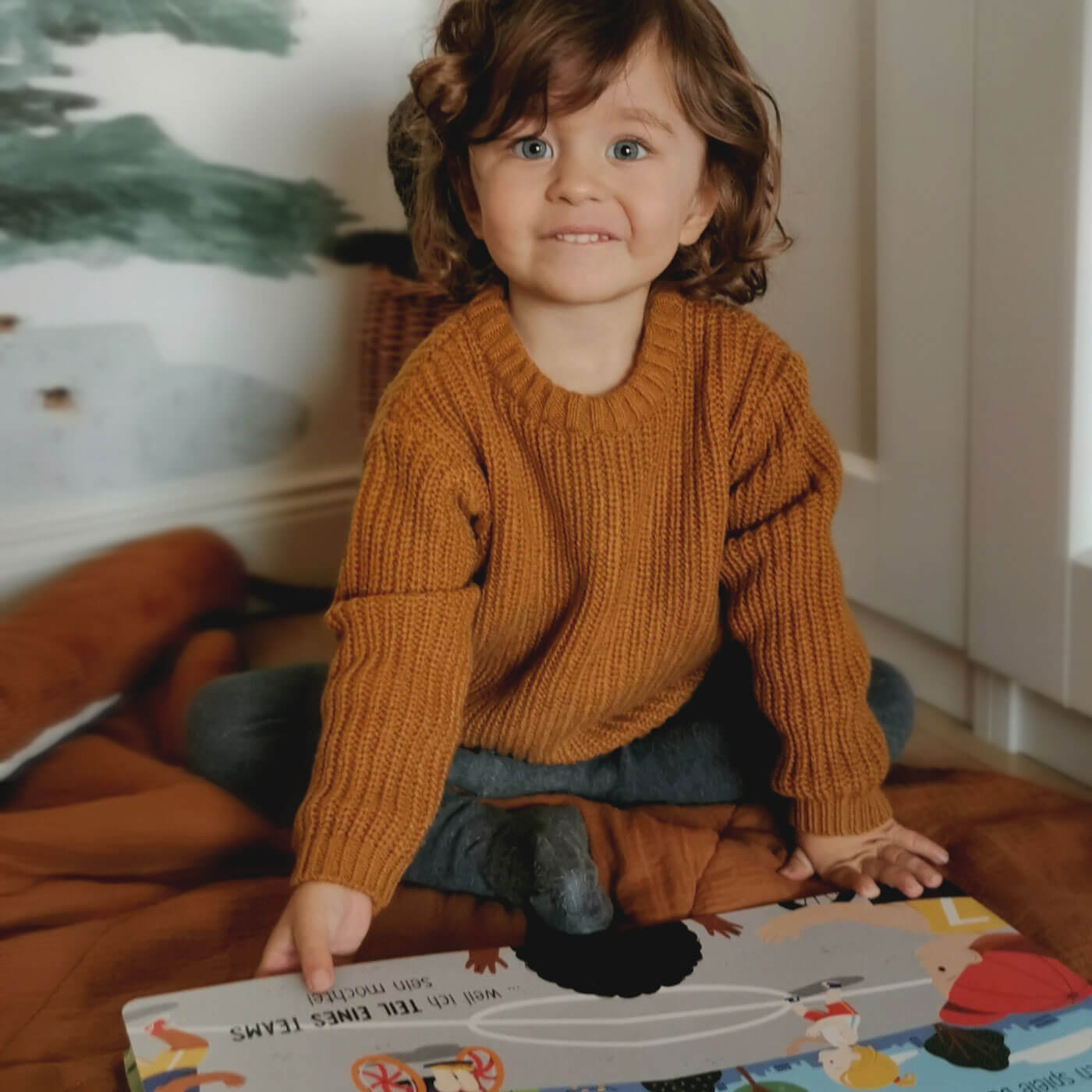 Ein kleines Kind mit lockigem braunem Haar und blauen Augen, das einen braunen Strickpullover trägt, sitzt im Schneidersitz auf einer Decke und lächelt in die Kamera, während es das Buch "Spielzeug ist für alle da & Kleidung ist für alle da" vom Zuckersüß Verlag in der Hand hält.