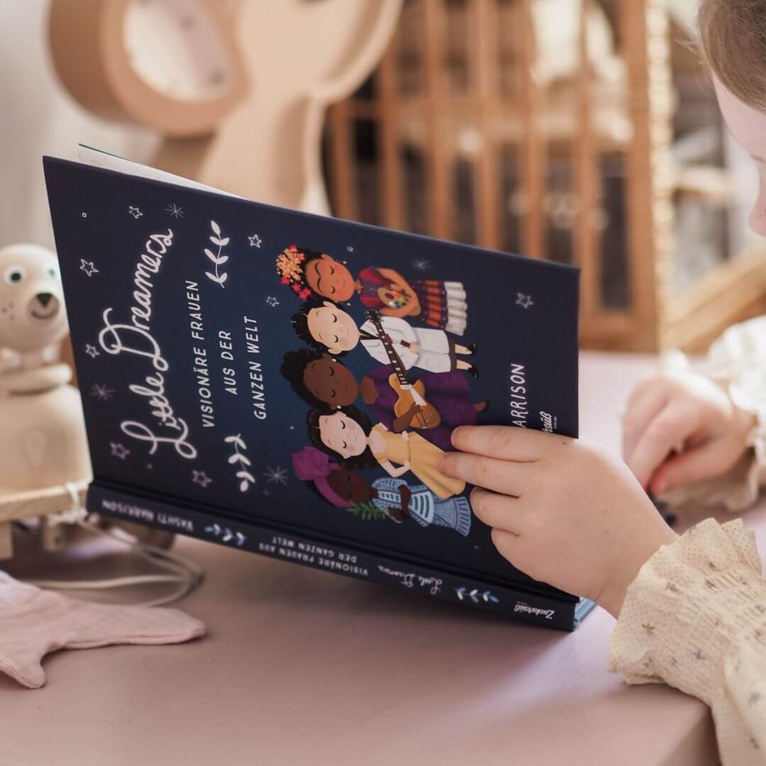 Ein Kind sitzt am pinken Magic Dreamer Set vom Zuckersüß Verlag und liest fasziniert "Little Dreamers: Visionäre Frauen aus der ganzen Welt". Ein Kuscheltier sowie schöne Deko sorgen für gemütliche Stimmung.