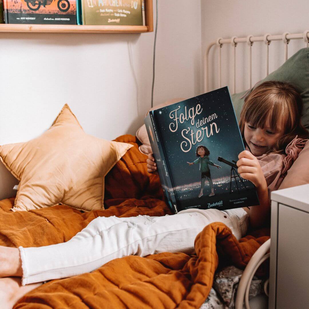Ein kleines Kind sitzt auf einem kuscheligen Bett mit einer senfgelben Decke und liest das Magic Dreamer Set vom Zuckersüß Verlag. Das illustrierte Cover zeigt ein Kind mit einem Teleskop unter dem Sternenhimmel, das zum Träumen und zur Neugier anregt.