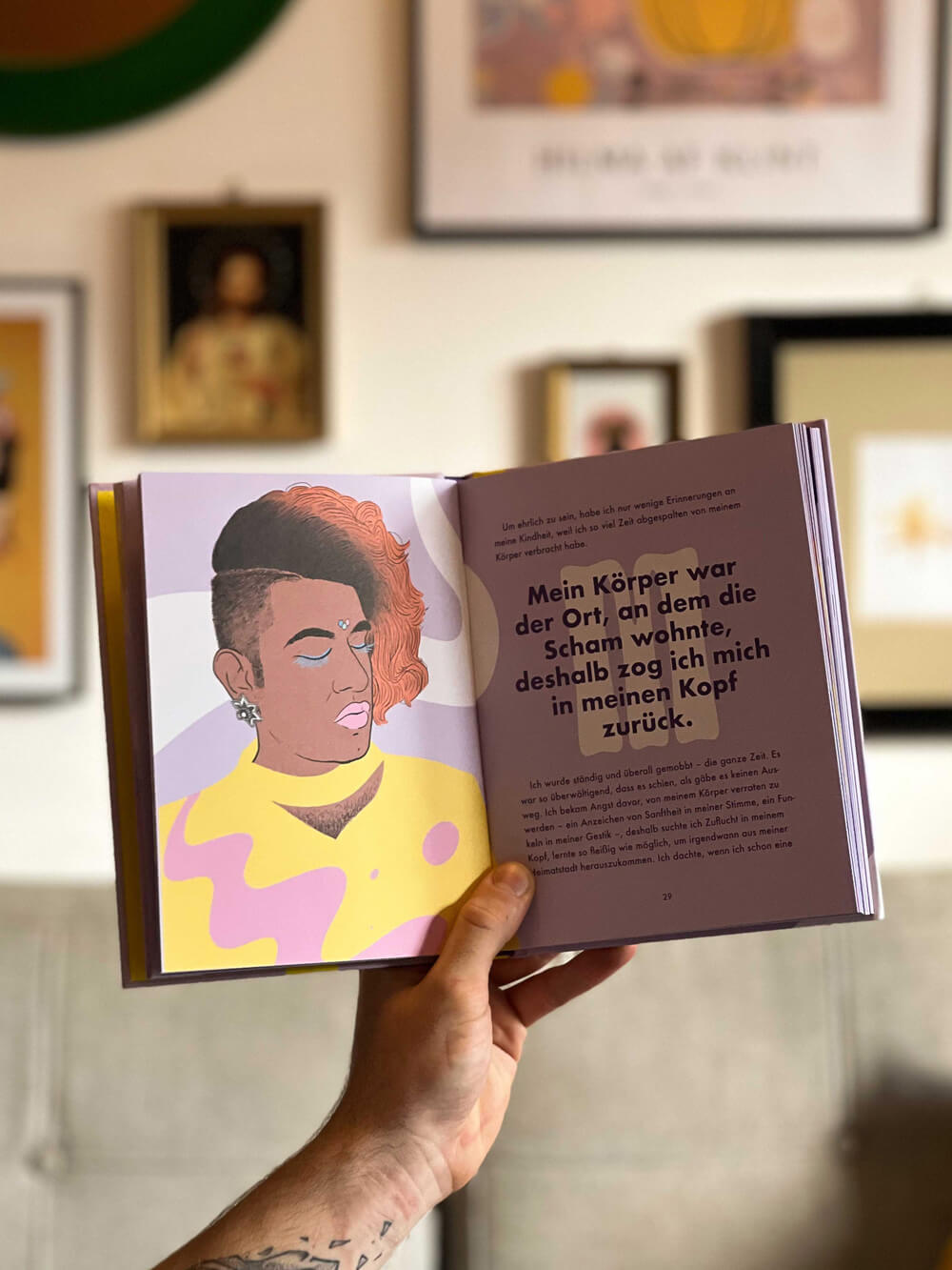 Eine Hand hält das Buch "Mehr als binär" von Alok Vaid-Menon auf, das links die Illustration einer kurzhaarigen Person mit Ohrringen und rechts einen Text über Identität jenseits des binären Geschlechtssystems zeigt. Die Kunst verschwimmt im Hintergrund.