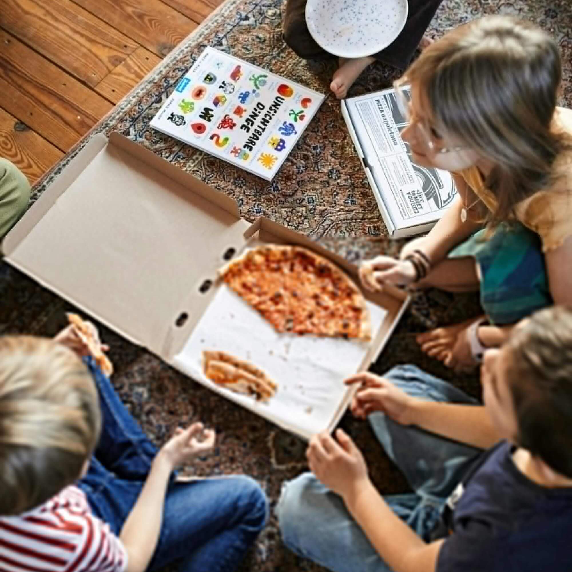 Vier Kinder sitzen auf einem gemusterten Teppich und essen gemeinsam Pizza aus einem offenen Karton. In der Mitte liegt das Cover vom Bilderbuch "Unsichtbare Dinge" von Andy J. Pizza und Sophie Miller. Daneben sind weitere Pizzakartons und ein leerer Teller zu sehen.
