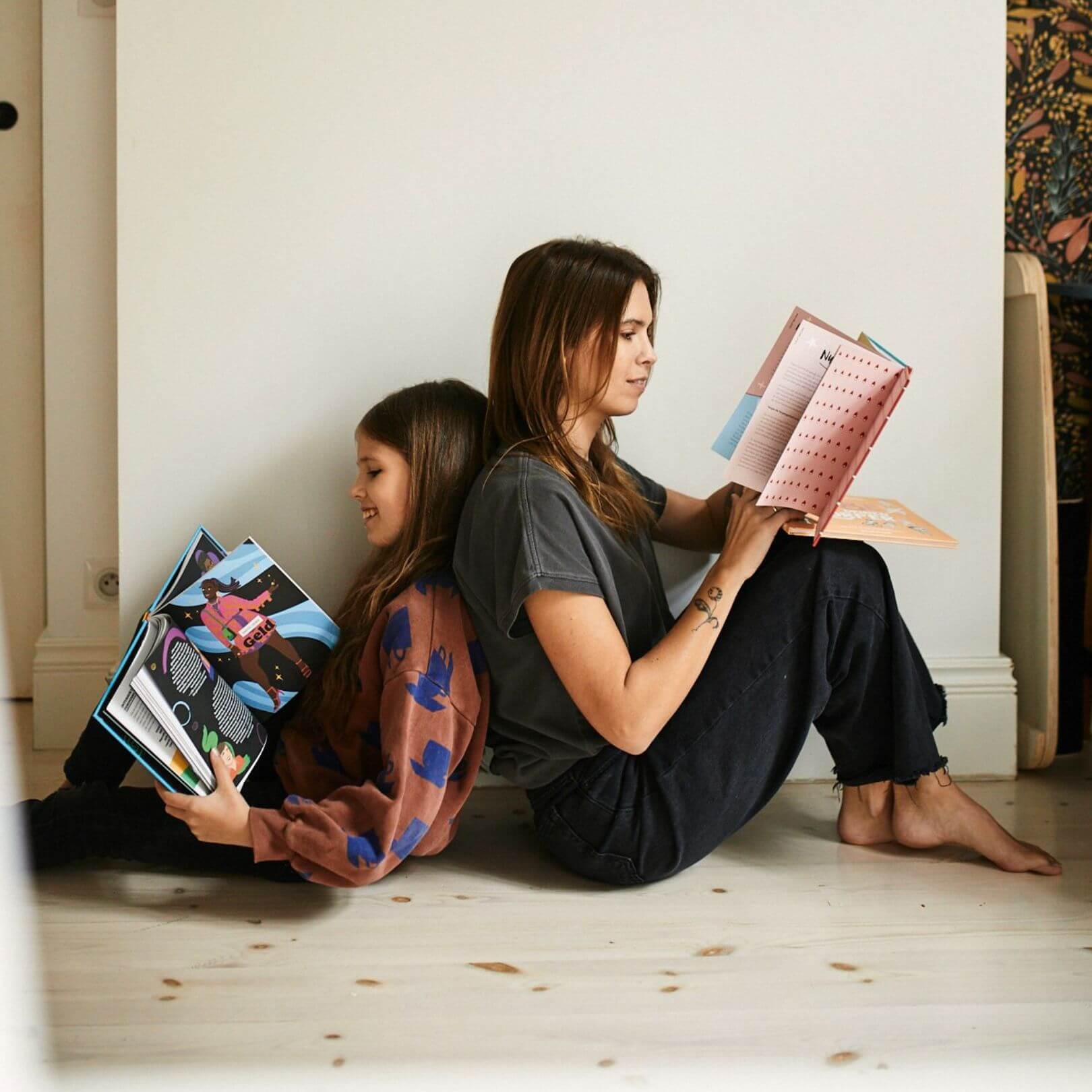 Eine Frau und ein junges Mädchen sitzen Rücken an Rücken auf einem Holzboden und lesen Bücher aus dem Young Feminist Starter Pack des Zuckersüß Verlags. 