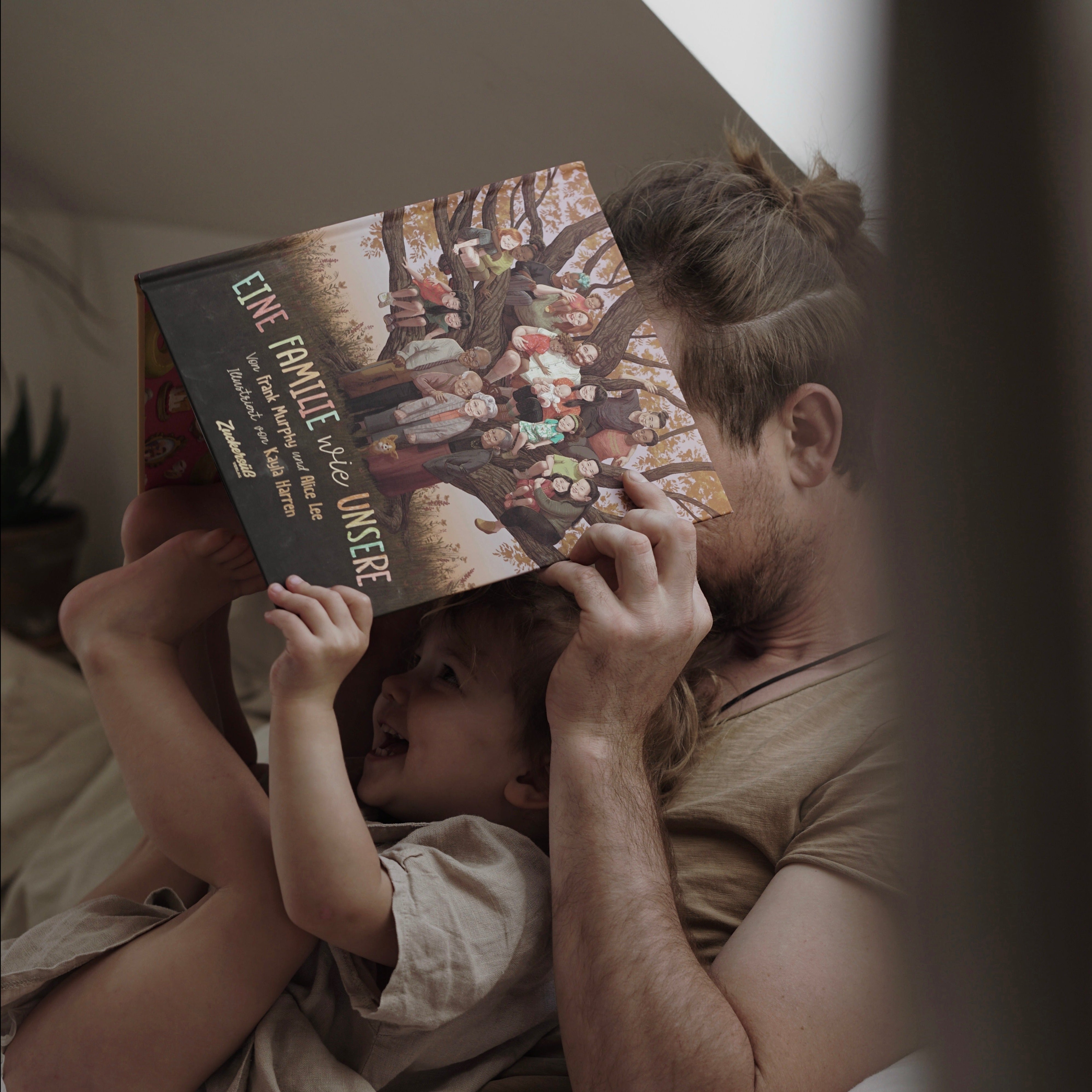 Eine erwachsene Person liegt mit einem Kind im Bett, beide lächeln, während sie gemeinsam das Buch „Eine Familie wie unsere“ anschauen. Die Szene strahlt Nähe, Geborgenheit und Freude am Vorlesen aus.