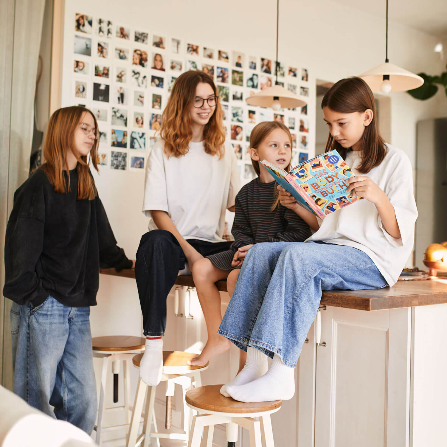 Vier Mädchen in Freizeitkleidung versammeln sich um eine Kücheninsel. Eine sitzt auf dem Tresen und liest aus dem bunten Pubertät Starter Pack des Zuckersüß Verlags, während die anderen zuhören und lächeln. Polaroidfotos erhellen die gemütliche Szene hinter ihnen.