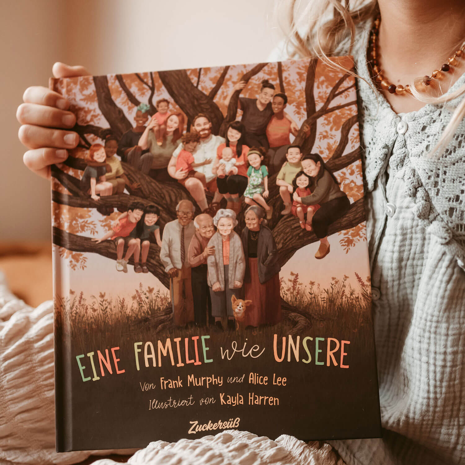Ein Kind hält das Buch "Eine Familie wie unsere" von Frank Murphy und Alice Lee in der Hand. Das Cover zeigt eine Gruppe von Menschen, die unter einem großen Baum stehen und Familie und Vielfalt feiern.