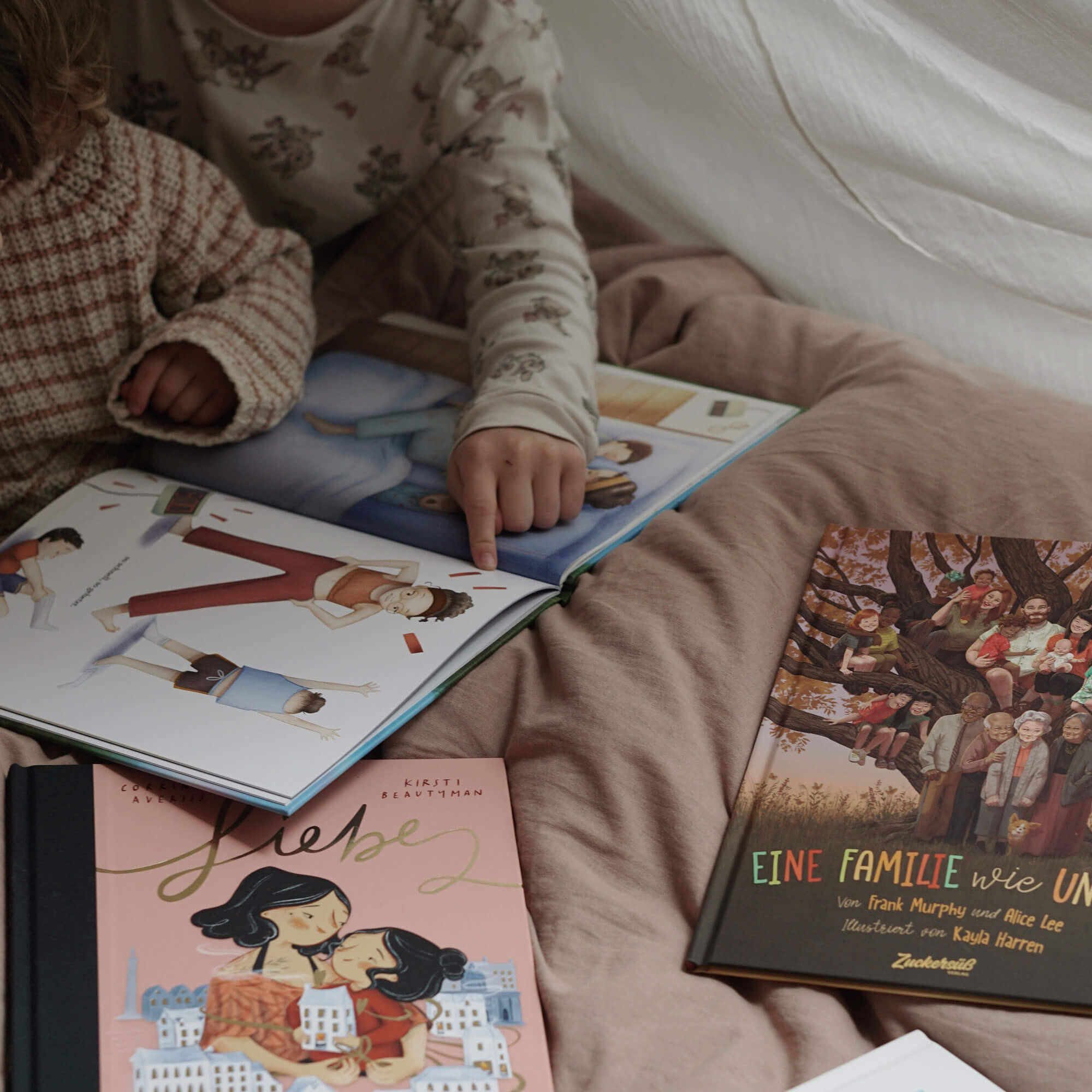 Zwei Kinder sitzen auf einem Bett und blättern gemeinsam in einem Bilderbuch aus dem „Das Richtig-Gute-Nacht-Paket“ vom Zuckersüß Verlag. Eines der Kinder zeigt auf eine Illustration im Buch. Um sie herum liegen weitere Titel wie „Liebe“ und „Eine Familie wie unsere“, die mit ihren warmen Covern eine gemütliche und geborgene Atmosphäre schaffen.