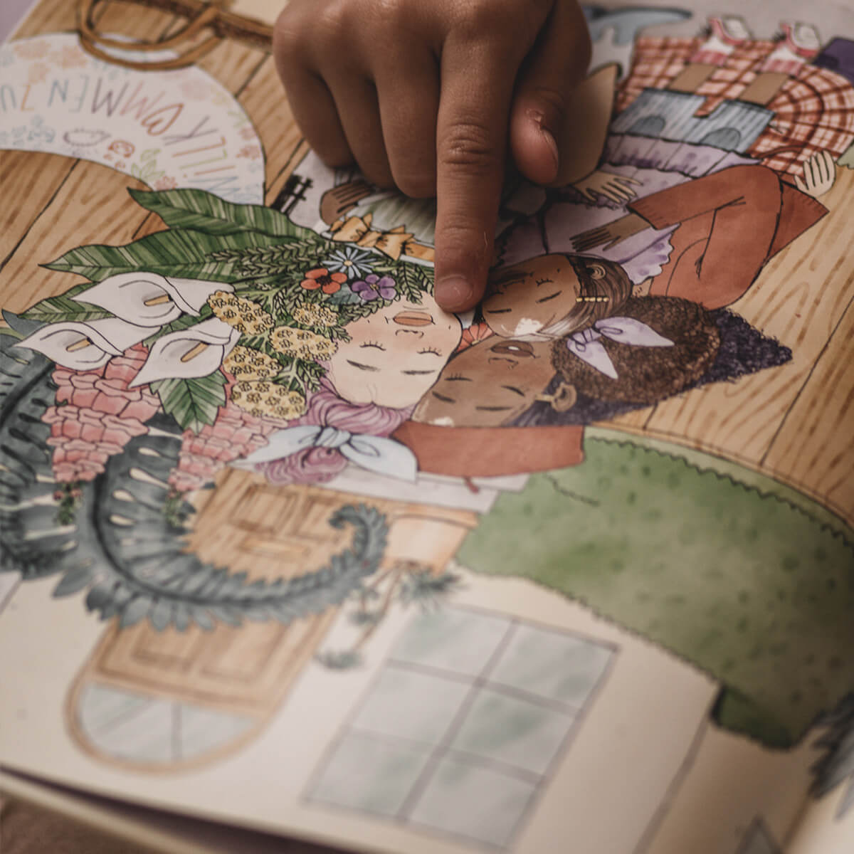 Die Hand eines Kindes zeigt auf eine Illustration in "Mama und Mami und ich: Die große Vermissung" von Nina LaCour, die zwei Menschen mit geschlossenen Augen zwischen Blumen in einem gemütlichen Zimmer zeigt.