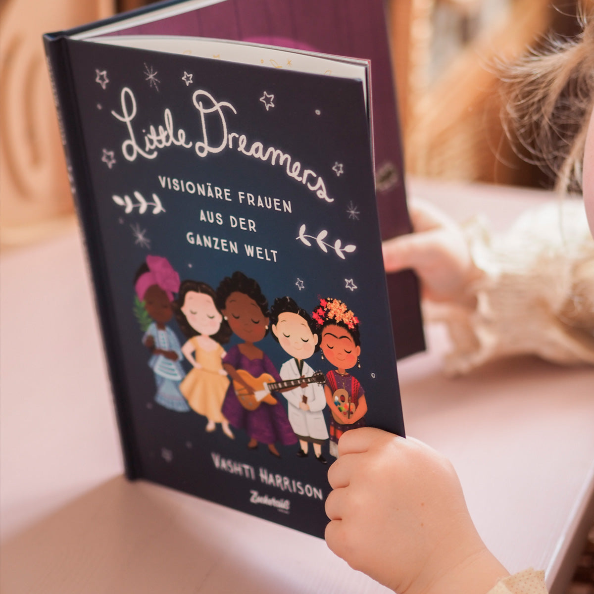 Auf dem Bild sieht man ein Kind, das das Buch „Little Dreamers" von Vashti Harrison in den Händen hält. Das Cover des Buches zeigt farbenfroh illustrierte Frauenfiguren aus unterschiedlichen Kulturen und Epochen, die für ihre inspirierenden Leistungen bekannt sind. Der Hintergrund ist dunkelblau mit Sternen und floralen Elementen.