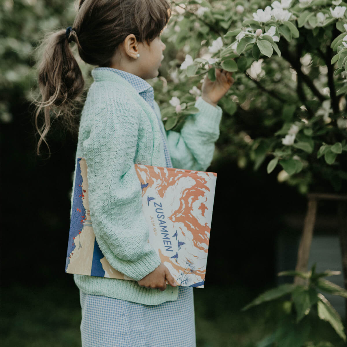 Ein Mädchen in mintgrünem Strickpullover und kariertem Kleid steht neben einem blühenden Baum und hält das Bilderbuch „Zusammen“ von Julia Kuo in der Hand.