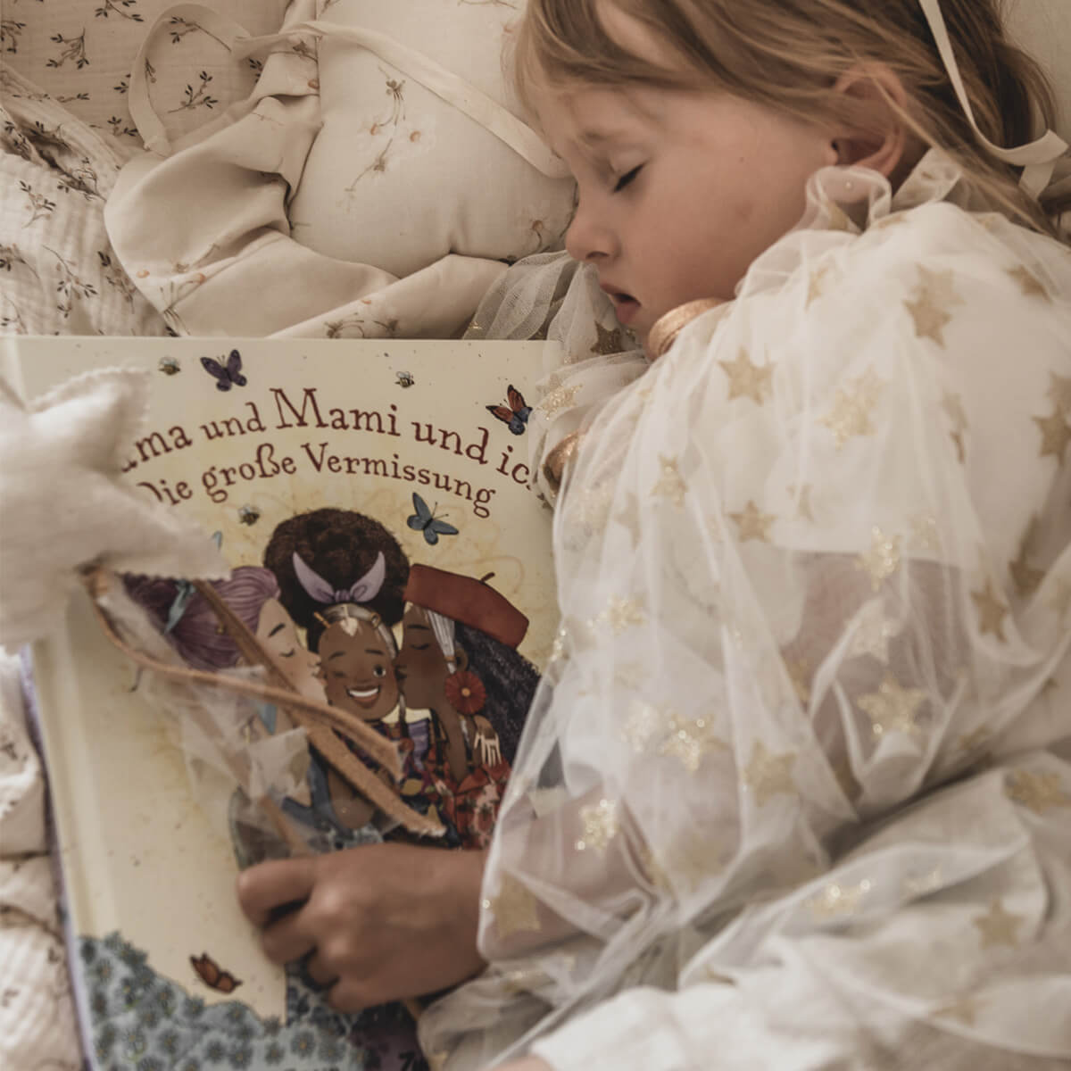 Ein kleines Kind in einem weißen Kleid mit goldenen Sternen schläft auf einer cremefarbenen Decke und hält das Bilderbuch „Die große Vermissung“ im Arm. Das Buch erzählt einfühlsam vom Vermissen einer nahestehenden erwachsenen Person und zeigt auf dem Cover eine liebevolle Umarmung.
