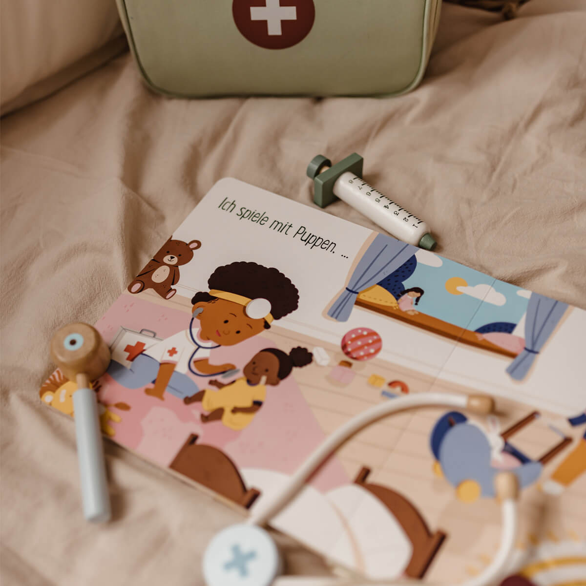 Eine deutsche Pappbilderbuchseite aus "Spielzeug ist für alle da!" von Susann Hoffmann zeigt ein Kind als Krankenschwester verkleidet, umgeben von medizinischen Spielzeugen wie einem Stethoskop und einer Spritze auf einer beigen Decke, mit einem grünen Verbandskasten im Hintergrund.