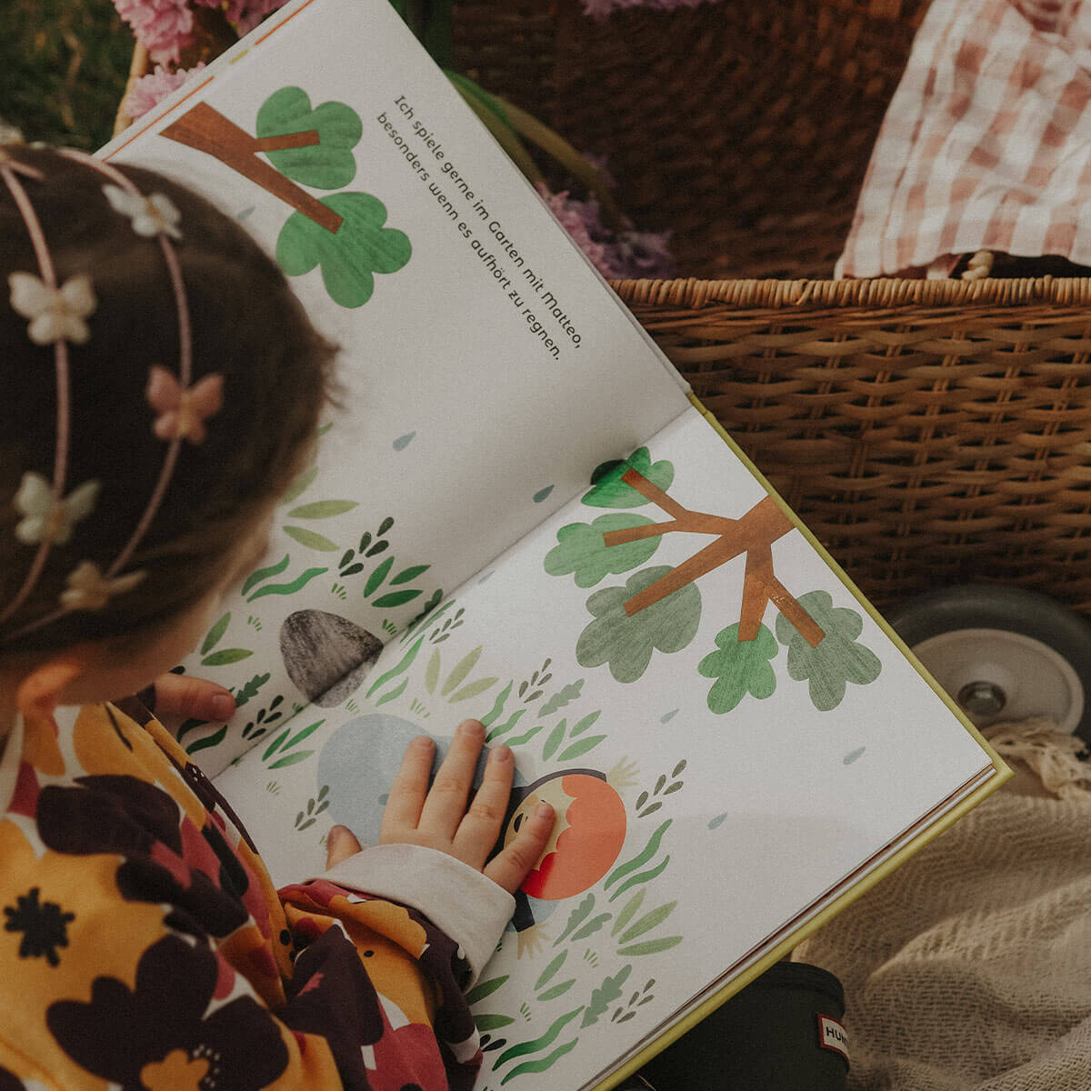 Ein kleines Kind mit einem Blumenstirnband liest im Freien neben einem Weidenkorb "Manchmal bin ich alles, was ich brauche" von Juliana Perdomo. Das illustrierte Buch zeigt einen Baum, Büsche und bunte Formen, die das Selbstvertrauen fördern sollen.