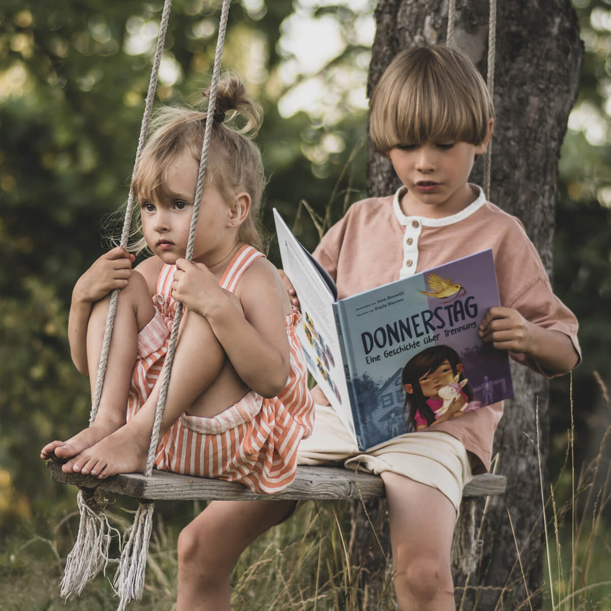 Zwei kleine Kinder sitzen auf einer Holzschaukel im Freien; das Mädchen in einem gestreiften Kleid umarmt ein Plüsch-Einhorn, während der Junge "Donnerstag" von Ann Bonwill liest. Bäume und Grün sorgen für eine friedliche Kulisse.