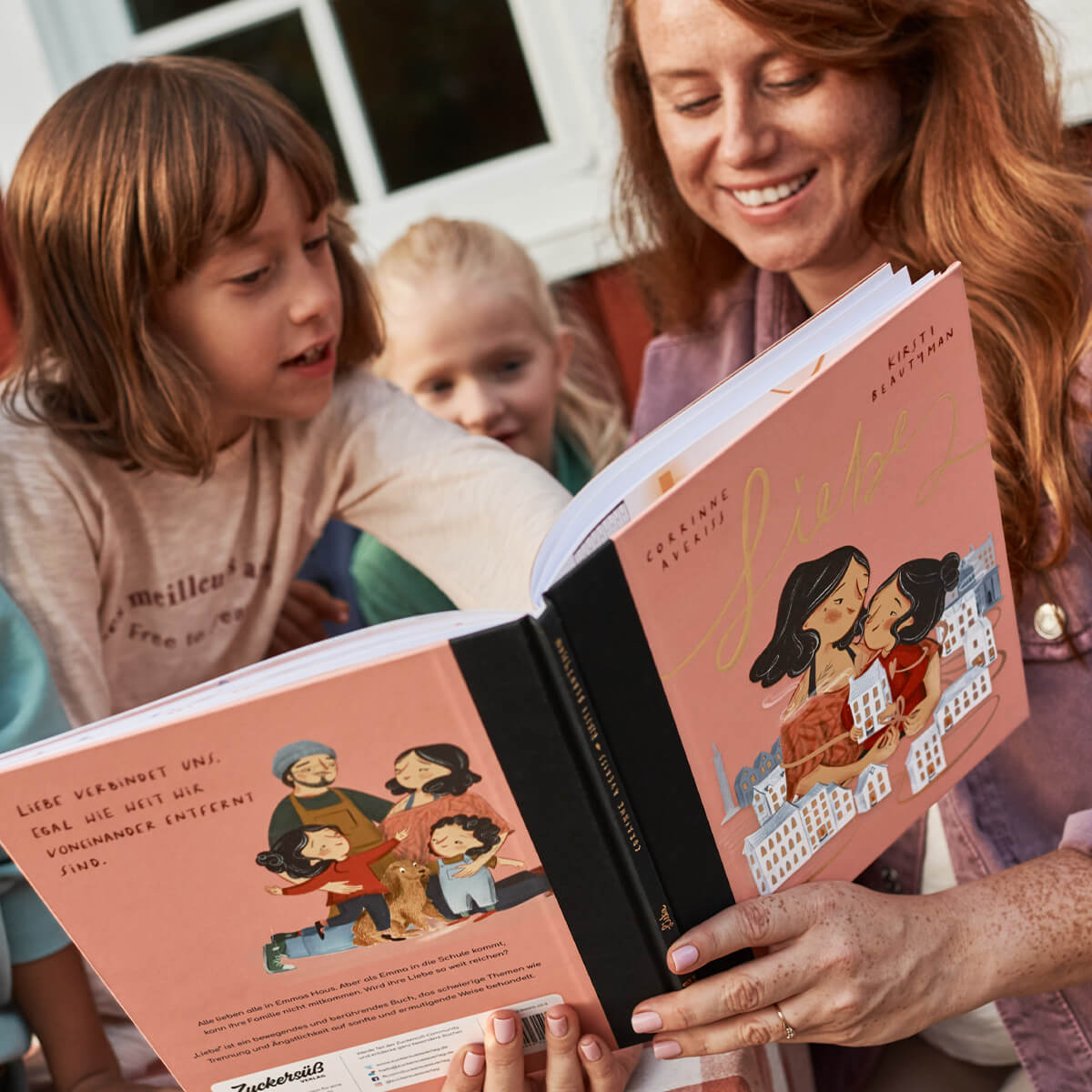 Eine erwachsene Person und drei Kinder sitzen im Freien und lesen „Mut & Liebe" vom Zuckersüß Verlag. Die Kinder schauen engagiert - eines zeigt auf das Buch -, die erwachsene Person lächelt warm, das Sonnenlicht erhellt den Moment, und die Kinder teilen ihre Gefühle miteinander.
