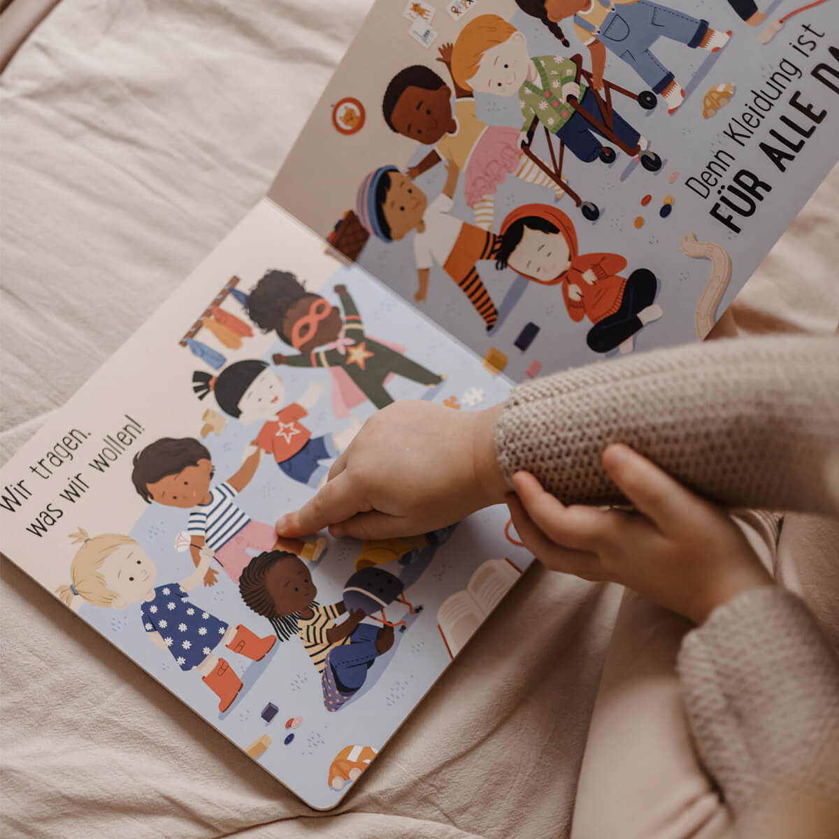 Ein Kind sitzt auf einem Bett und liest das Bilderbuch "Kleidung ist für alle da!" von Susann Hoffmann, das verschiedene Kinder in bunter Kleidung zeigt. Das Kind zeigt auf die Seite mit dem Text: Wir tragen, was wir wollen!.