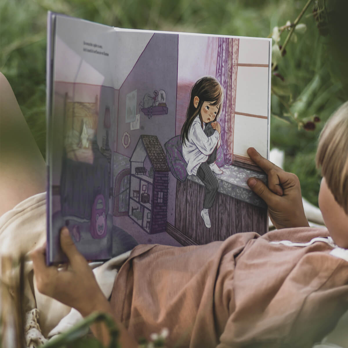 Ein Kind liest im Freien "Donnerstag" von Ann Bonwill und betrachtet die Illustration eines Mädchens in einem lila Zimmer auf einer Fensterbank, das ein Plüsch-Einhorn umarmt und nachdenklich dreinschaut.