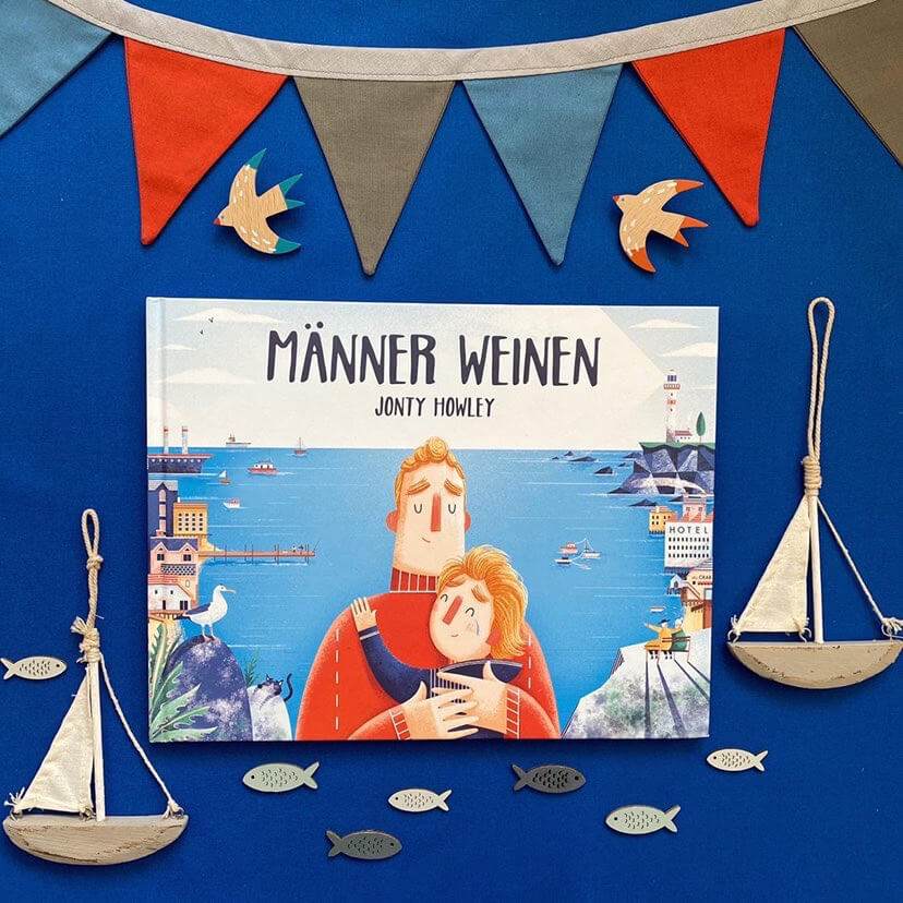 Das Kinderbuch "Männer weinen" von Jonty Howley liegt auf blauem Hintergrund, umgeben von bunter Wimpelkette sowie maritimen Dekorationen.