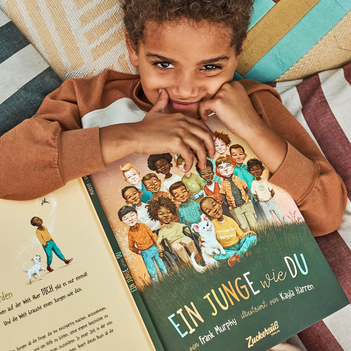 Ein lächelndes Kind mit lockigem Haar liegt auf einer gestreiften Decke und hält das Frank-Murphy-Buch "Ein Junge wie du" in der Hand, das auf dem Cover verschiedene Kinder und einen Hund zeigt.