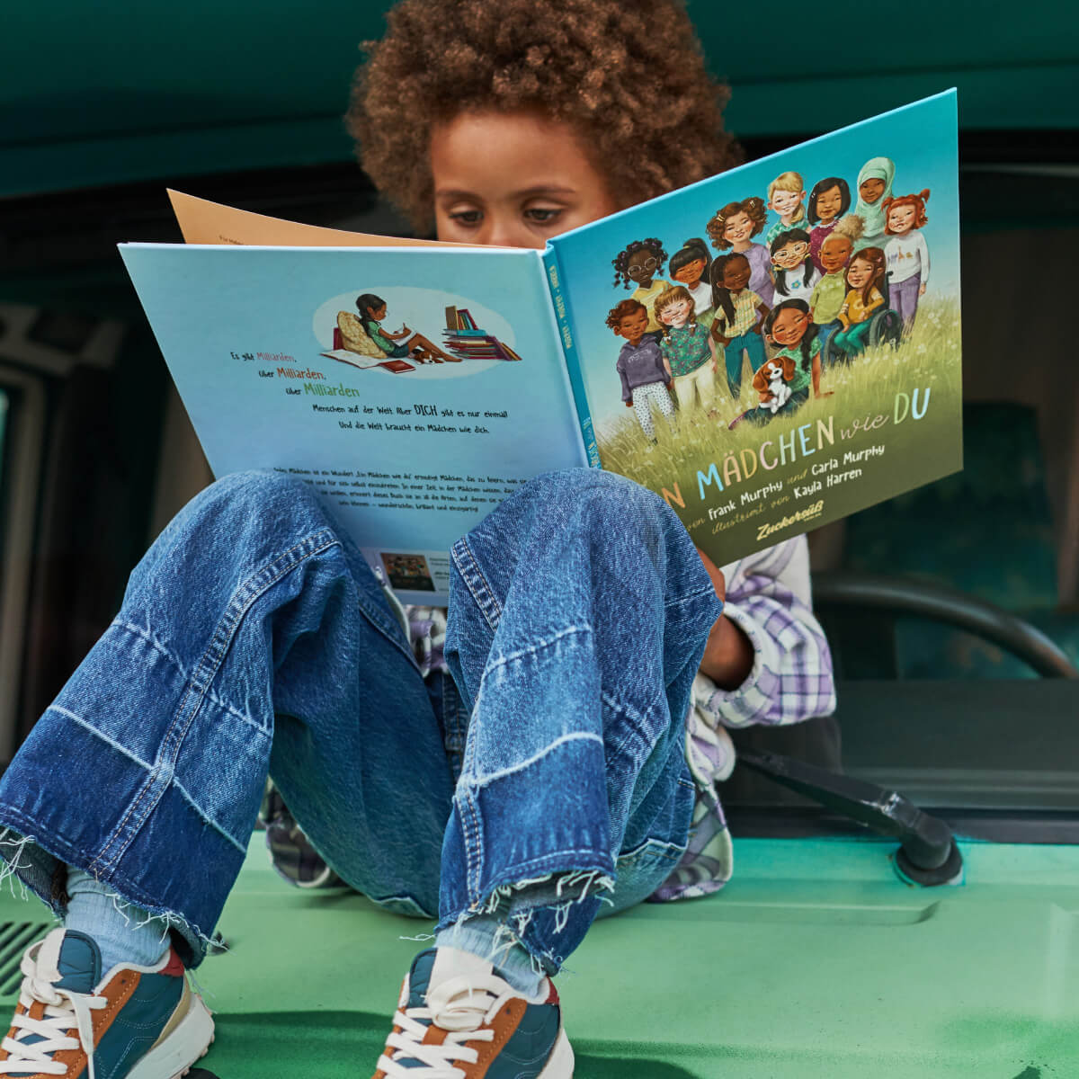 Ein Kind mit lockigem Haar sitzt mit angewinkelten Beinen auf einer grünen Fläche, trägt Patchwork-Jeans und bunte Sneaker und liest konzentriert das Kinderbuch „Ein Mädchen wie du“ von Frank und Carla Murphy.