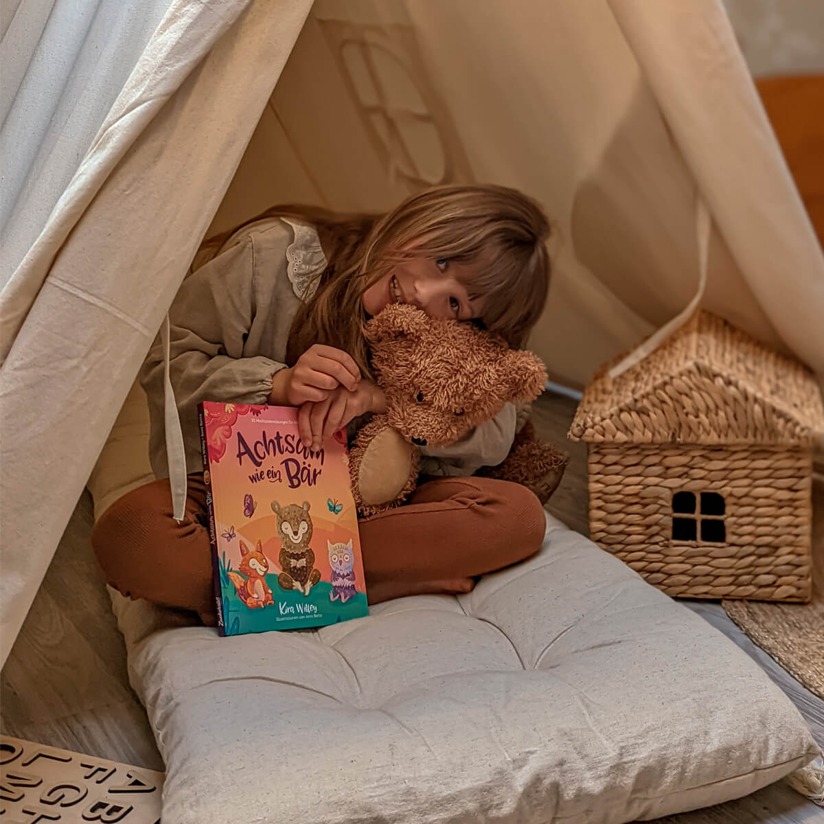 Ein Kind sitzt gemütlich in einem beigen Spielzelt auf einem weichen Kissen, kuschelt sich an einen braunen Teddybär und hält das bunte Kinderbuch „Achtsam wie ein Bär“ von Kira Willey in der Hand. Neben dem Kind steht ein kleines geflochtenes Spielhaus. Die Szene wirkt warm und geborgen.