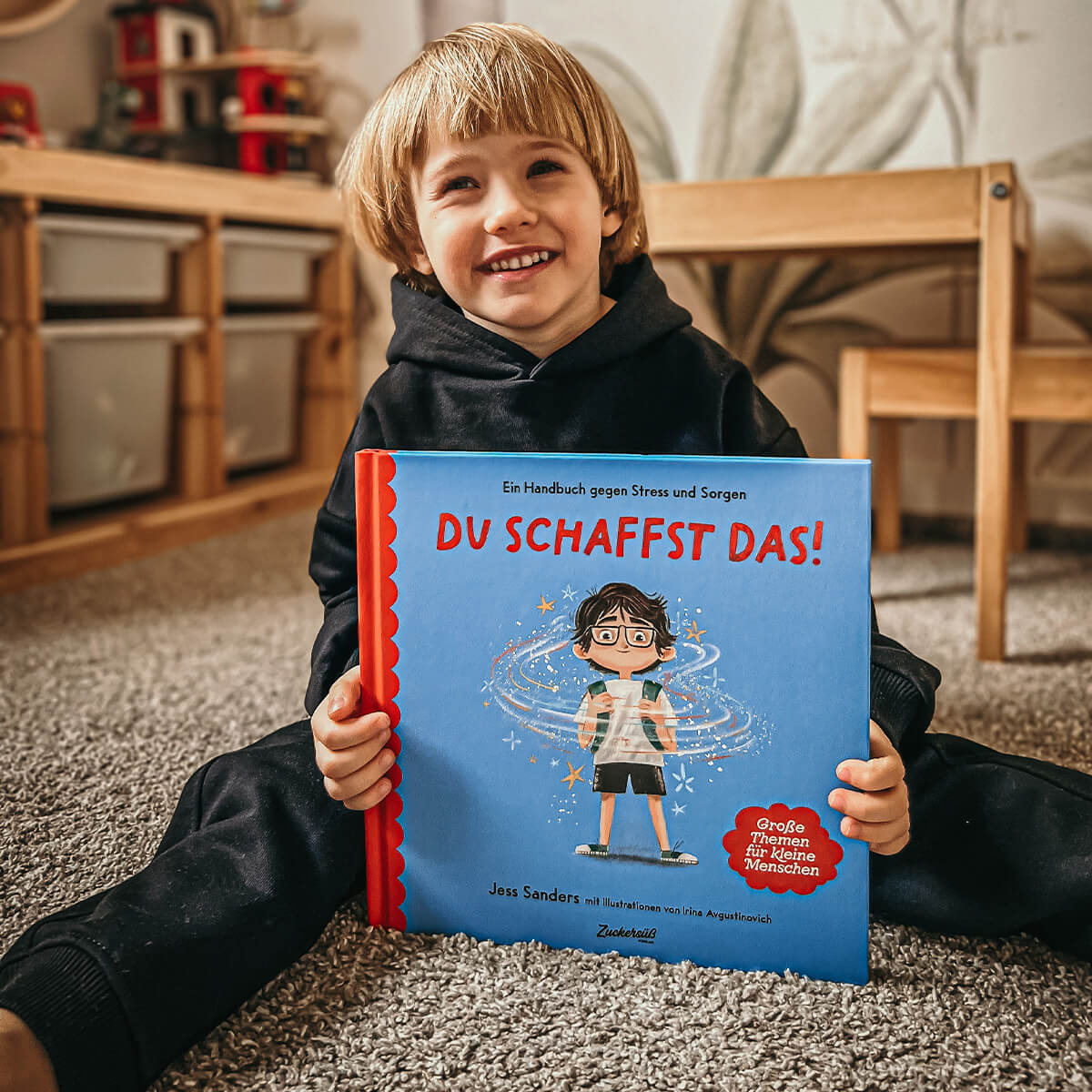 Ein lächelndes Kind in einem schwarzen Kapuzenpulli sitzt auf einem Teppichboden und hält das Buch 