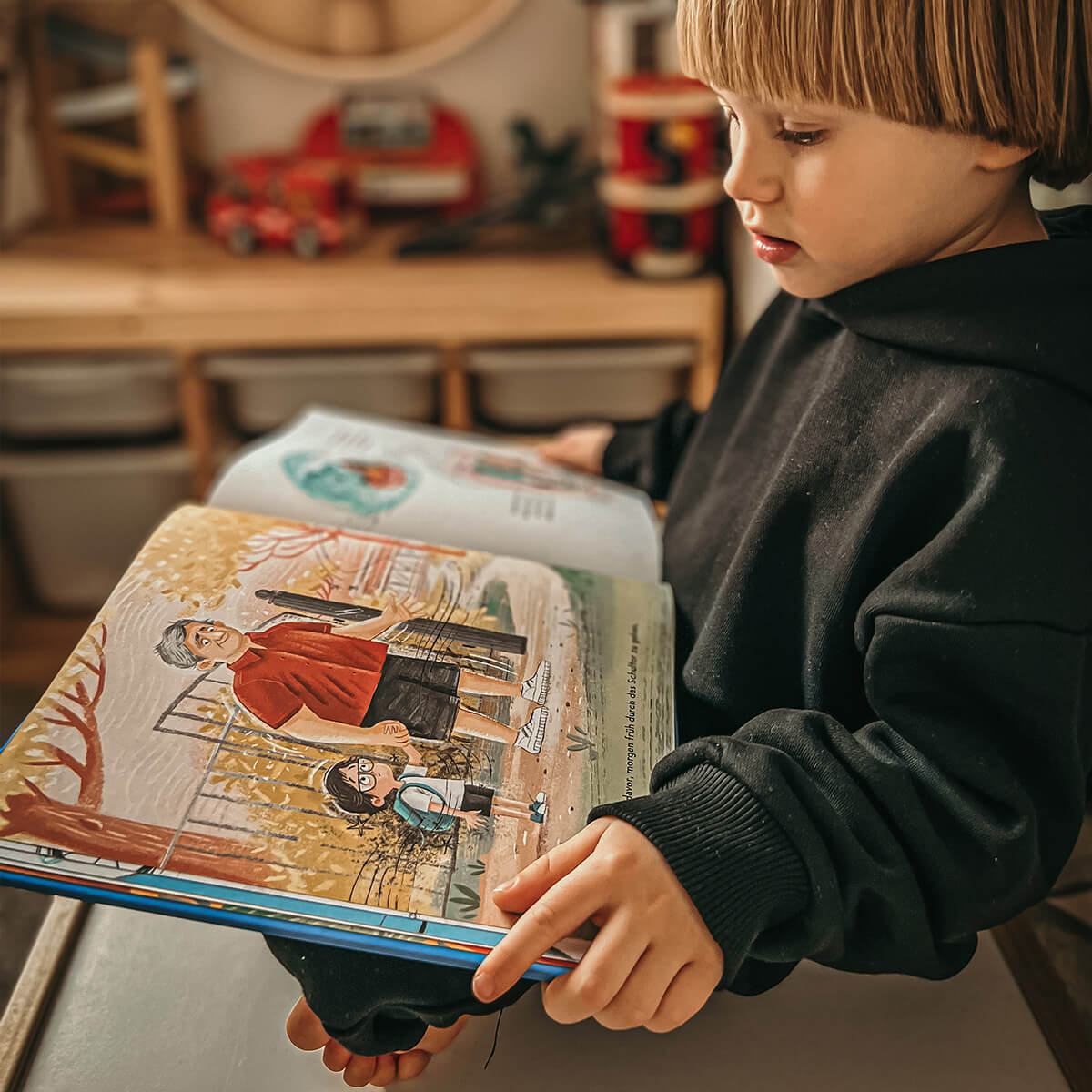 Ein Kind in einem schwarzen Kapuzenpulli sitzt in einem Haus und liest das Buch "Du schaffst das!" von Jessica Sanders. Die aufgeschlagenen Seiten zeigen eine bunte Illustration eines Erwachsenen und eines Kindes, die draußen zwischen Herbstblättern stehen.