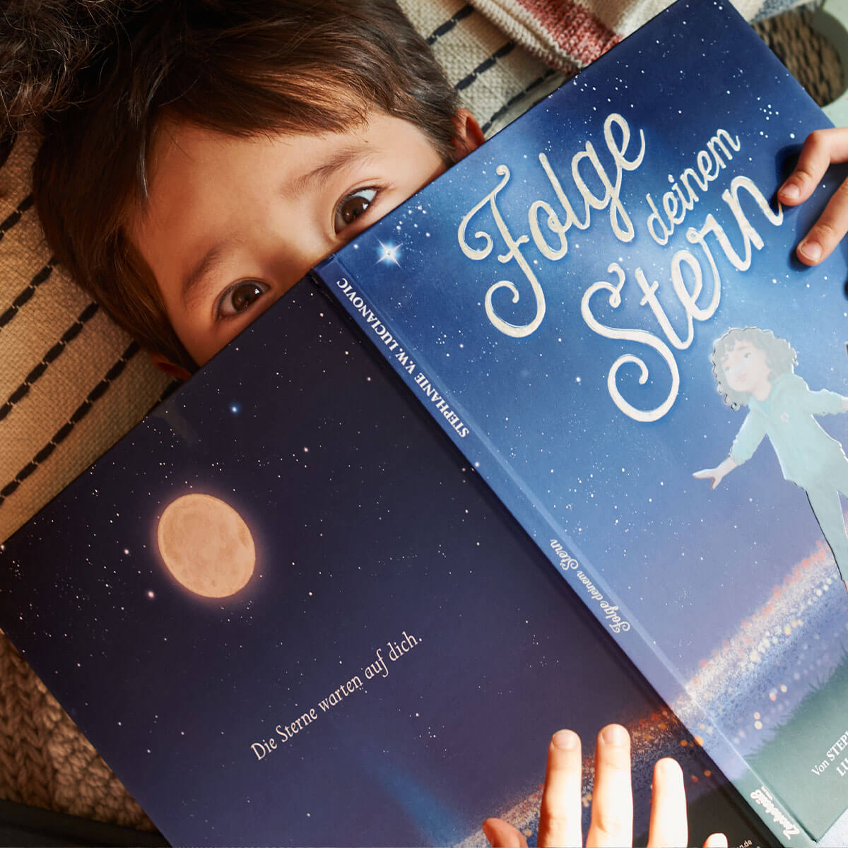 Ein Kind auf einer gestreiften Decke hält das aufgeschlagene Kinderbuch "Folge deinem Stern" von Stephanie V.W. Lucianovic in der Hand, dessen sternförmiges Cover junge Leser zum Träumen von Astronomie und Weltraum anregt.