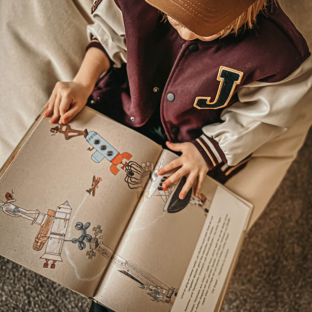 Ein Kind in einer kastanienbraunen und cremefarbenen Jacke mit einem gelben J-Aufnäher sitzt vertieft in Kurz und Klein von Alastair Chisholm und studiert die farbenfrohen Illustrationen und den spannenden Text über Höhlenmenschen und ihre frühen Erfindungen.