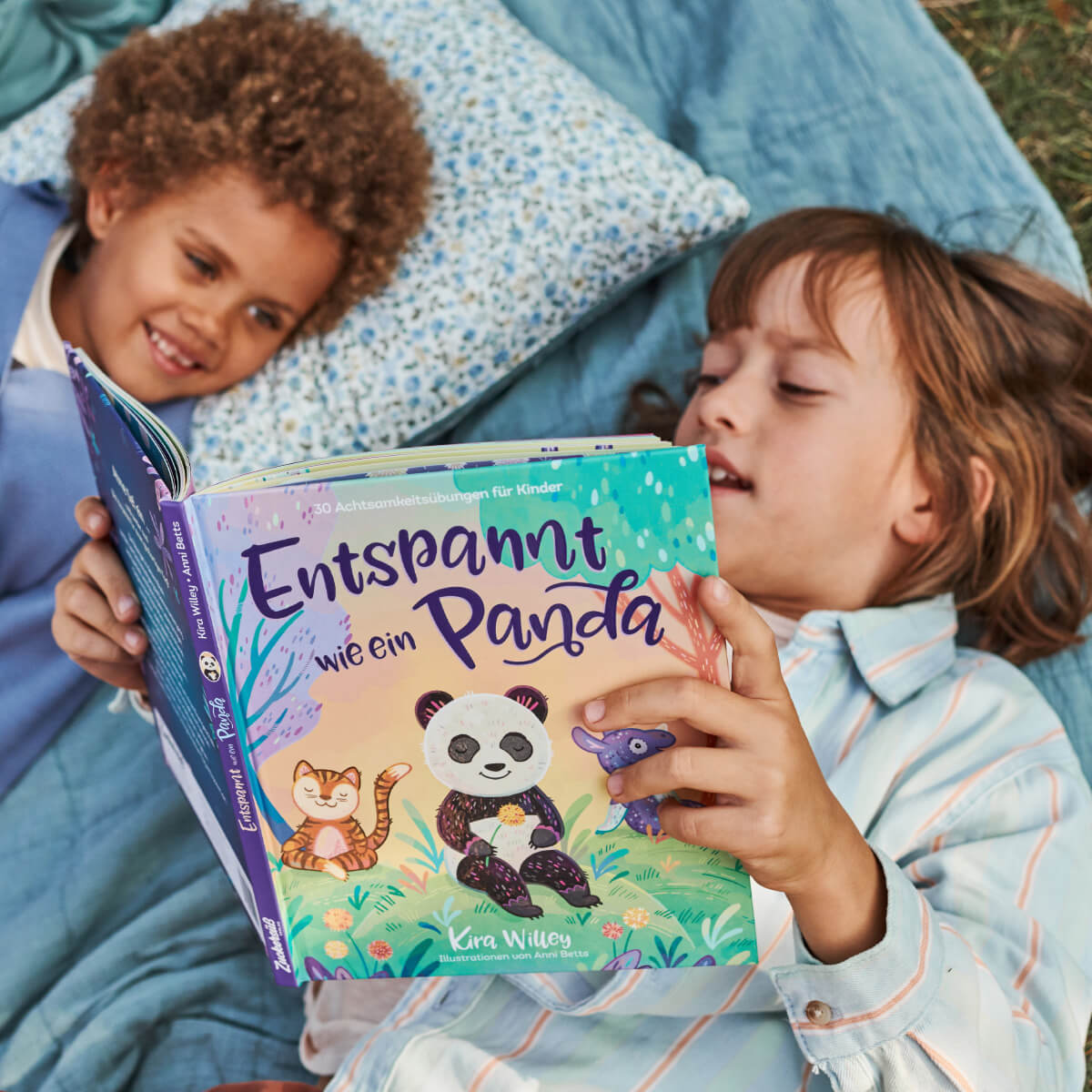 Zwei Kinder liegen auf einer Decke im Freien, eines hält das bunte Buch „Entspannt wie ein Panda“ von Kira Willey in den Händen. Beide lächeln, während sie gemeinsam lesen.