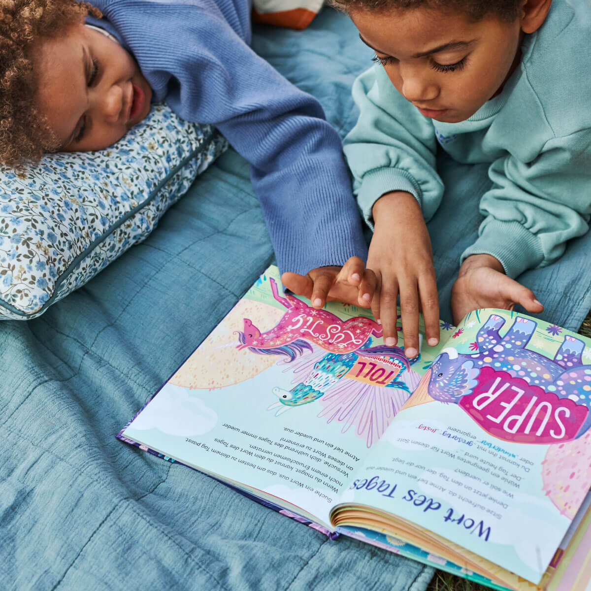 Zwei Kinder liegen auf einem Bett mit blauer Bettwäsche und einem geblümten Kissen und lesen 