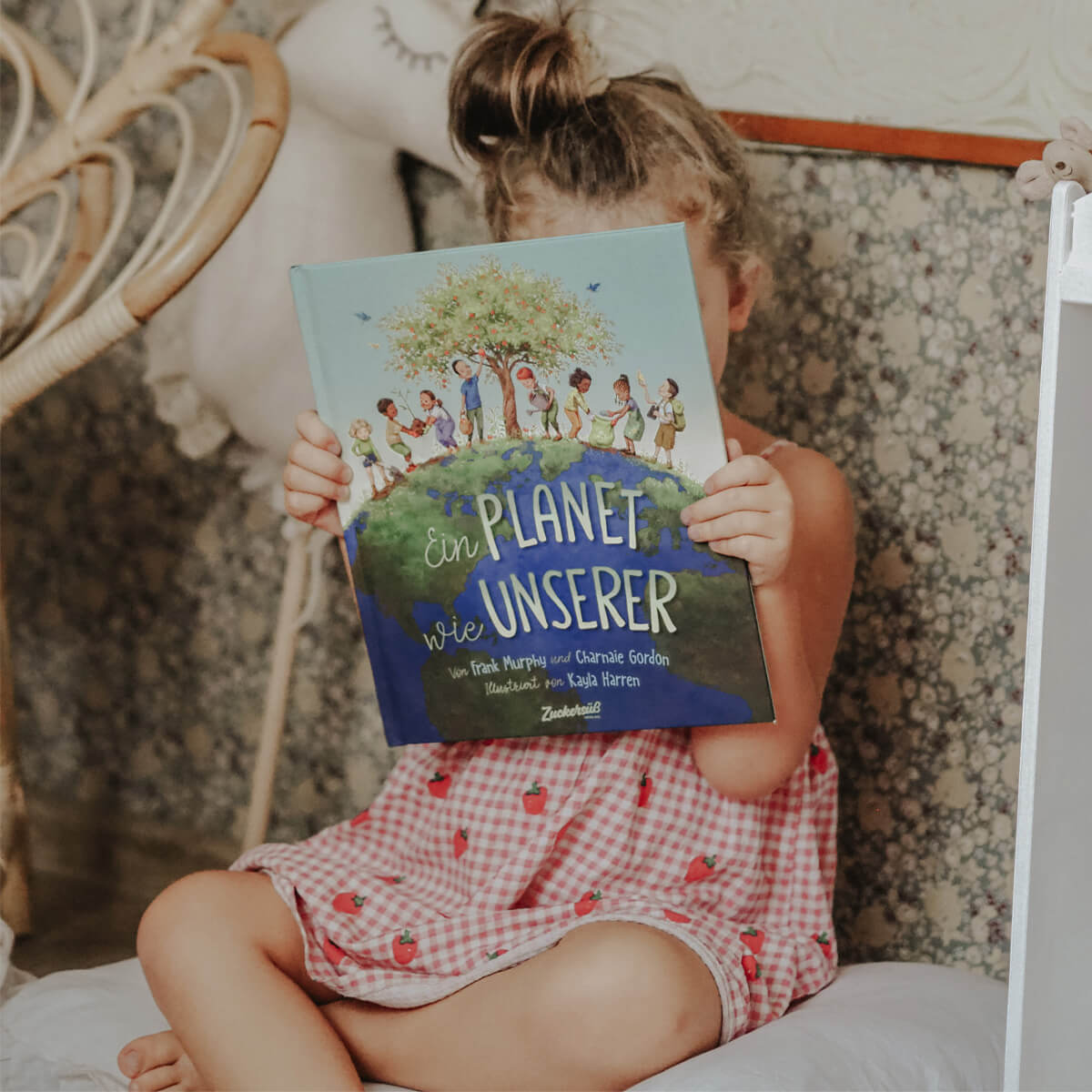 Ein kleines Kind in einem rot-weiß karierten Kleid sitzt auf einem Bett und hält das Buch „Ein Planet wie unserer“ aus dem Starterpaket „Welt verändern“ des Zuckersüß Verlags vor das Gesicht. Auf dem Cover sind diverse Kinder zu sehen, die sich auf einem grünen Hügel bei einem großen Baum an den Händen halten – ein Bild, das Gemeinschaft, Vielfalt und Verantwortung für unseren Planeten vermittelt.