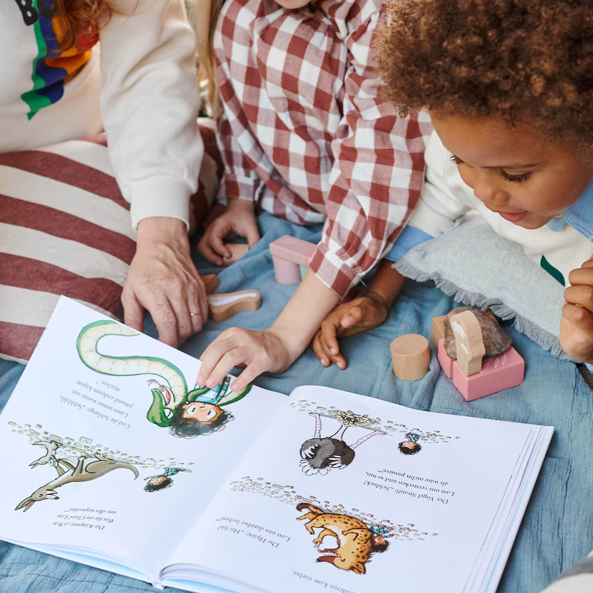 Drei Kinder sitzen eng beieinander auf einer blauen Decke und betrachten die herzerwärmenden Illustrationen im aufgeschlagenen Bilderbuch "Häschen tröstet" von Cori Doerrfeld. Daneben liegen hölzerne Tierspielzeuge; eines der Kinder trägt ein kariertes Hemd.