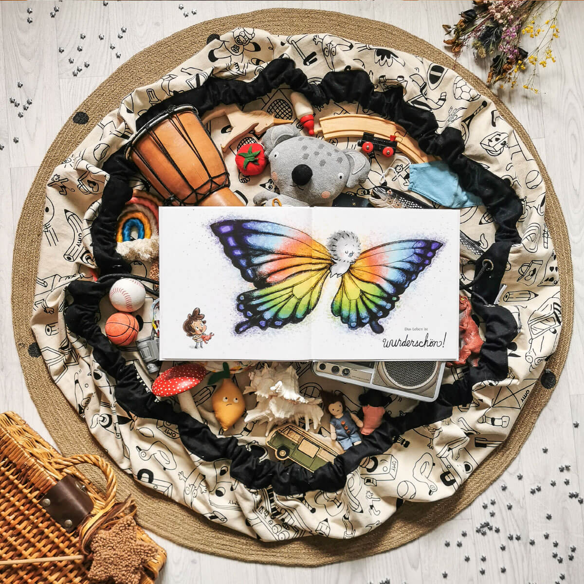 Auf einer Kinderspielmatte liegen Spielsachen und ein Exemplar des Buches "So ist das Leben" von Ame Dyckman, auf dem ein Kind mit leuchtenden Schmetterlingsflügeln abgebildet ist. In der Nähe laden ein Weidenkorb und Trockenblumen zu kleinen Abenteuern auf dem Holzboden ein.