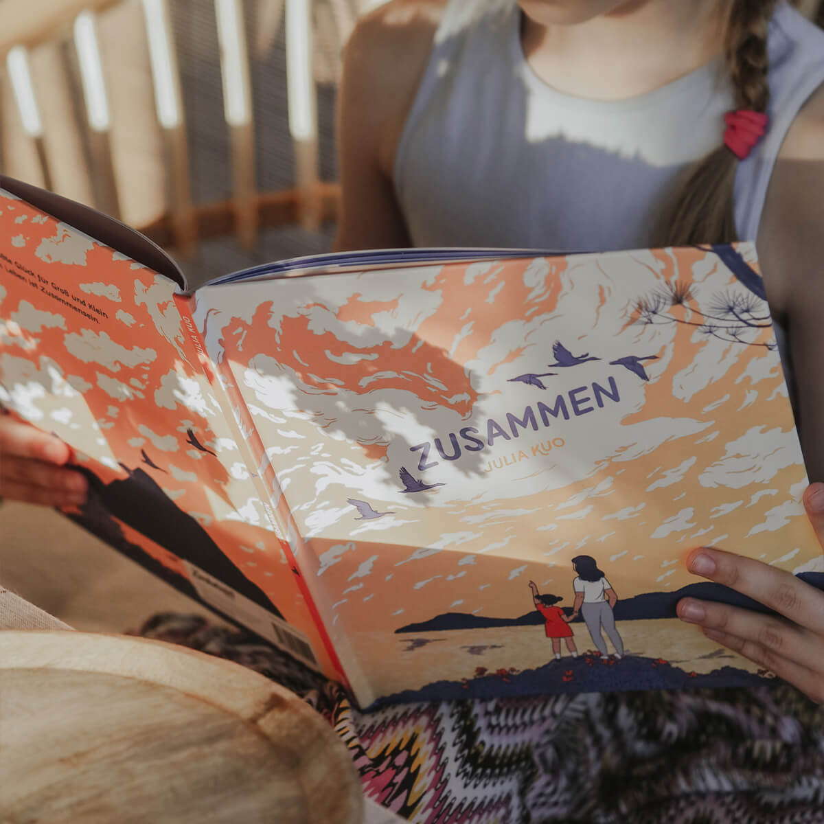 Ein Kind mit seitlichem Zopf sitzt auf einem gemusterten Kissen und hält das Bilderbuch Zusammen von Julia Kuo in den Händen. Auf dem Cover sind eine erwachsene Person und ein Kind zu sehen, die gemeinsam in einen orangefarbenen Himmel mit fliegenden Vögeln blicken.