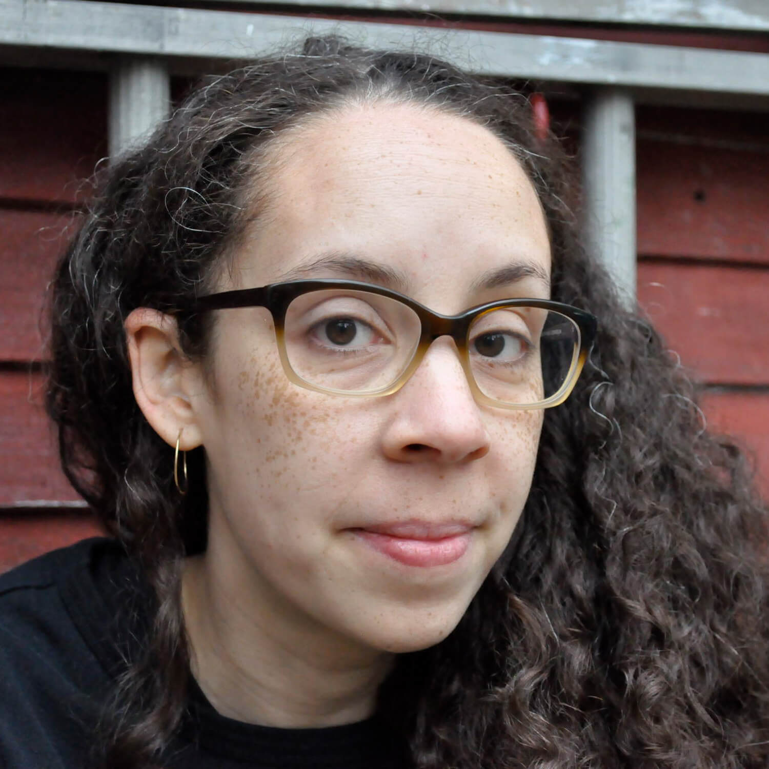 Mit lockigem braunem Haar, Brille und goldenen Ohrringen steht eine selbstbewusste Person vor einer roten Holzwand mit weißer Umrandung und hält "Das Buch vom Antirassismus" von Tiffany Jewell in der Hand, das den Stolz auf ihre soziale Identität widerspiegelt.