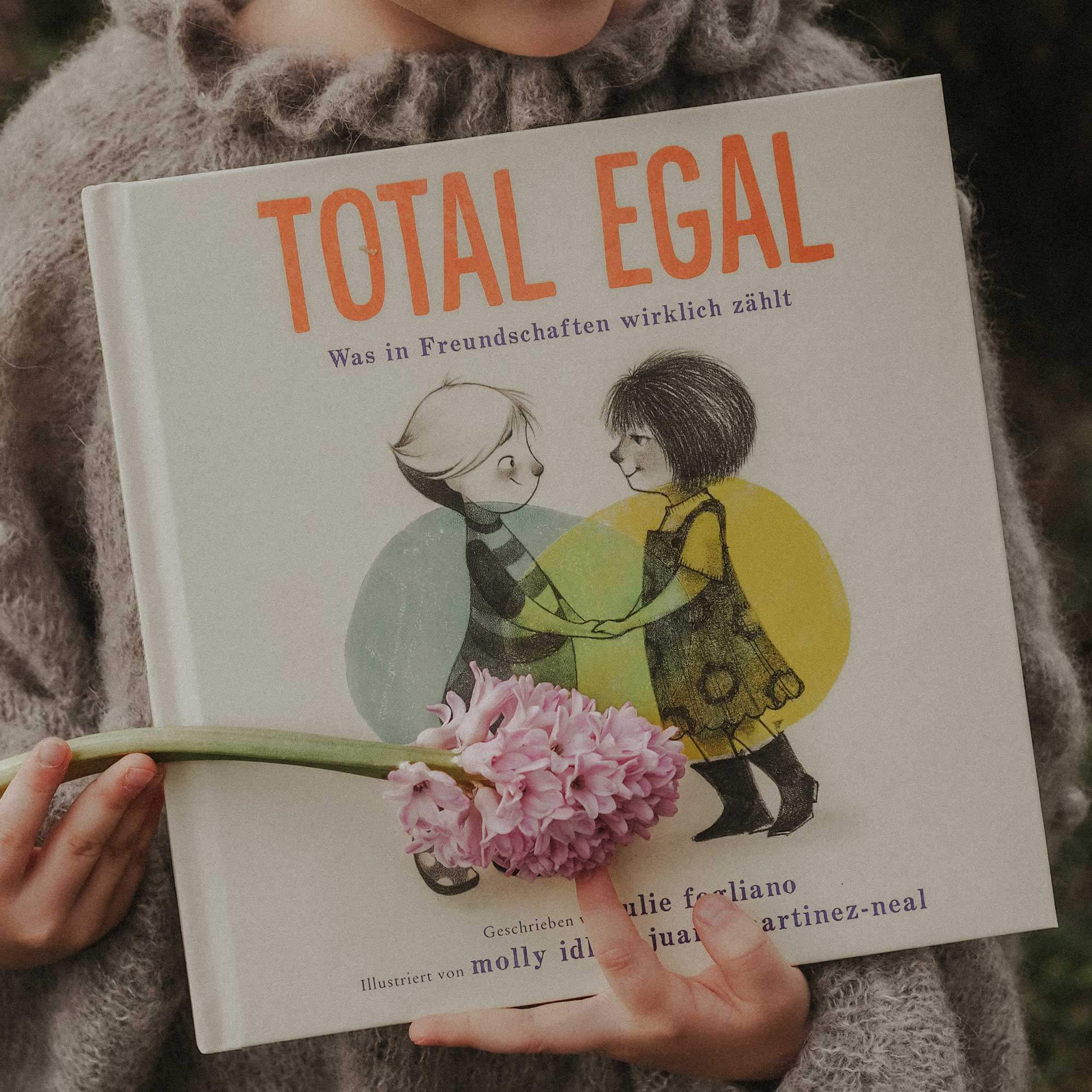 Ein Kind in einem grauen Pullover hält Julie Foglios "Total egal" in der Hand. Auf dem Cover sind zwei lächelnde Kinder zu sehen, die sich an den Händen halten. Eine Geschichte, die das Einfühlungsvermögen und die Essenz wahrer Freundschaft feiert.