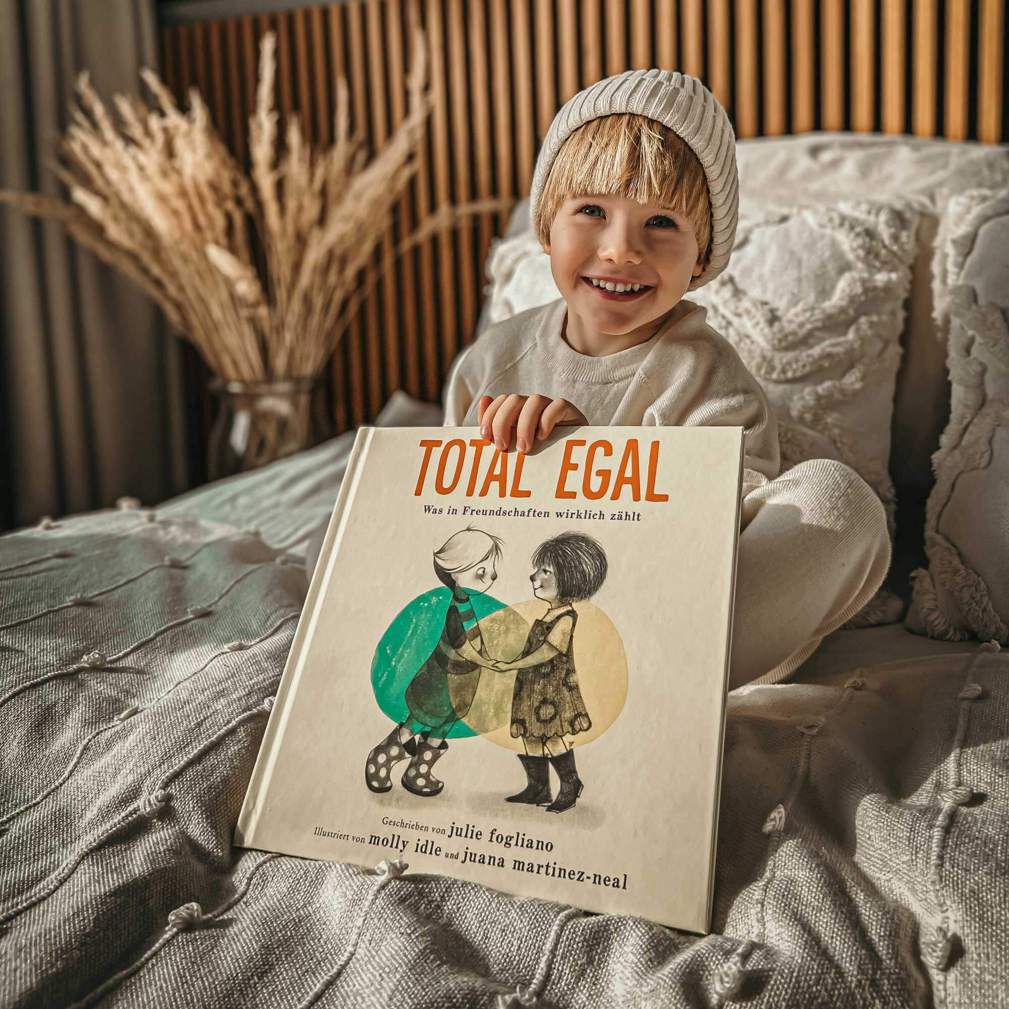 Ein lächelndes Kind in einem weißen Outfit und einer Mütze sitzt auf einem Bett und hält "Total egal" von Julie Fogliano in der Hand, das auf dem Cover illustrierte Kinder zeigt.